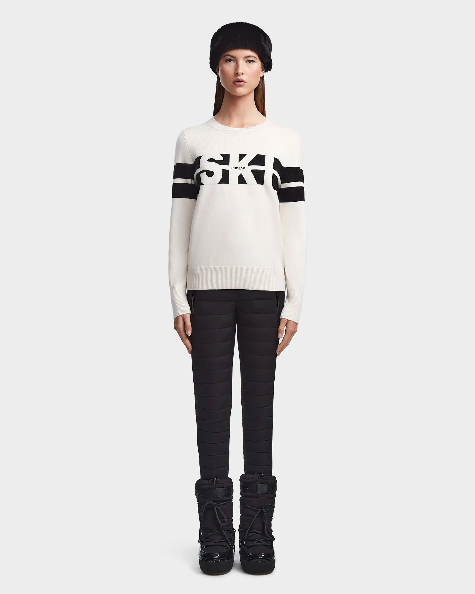 A woman faces forward wearing the ODESSA - 8523540 Iceland soft-knit Rudsak sweater with black sleeve stripes and SKI on the chest, paired with black quilted pants, snow boots, and a furry black hat for winter warmth.