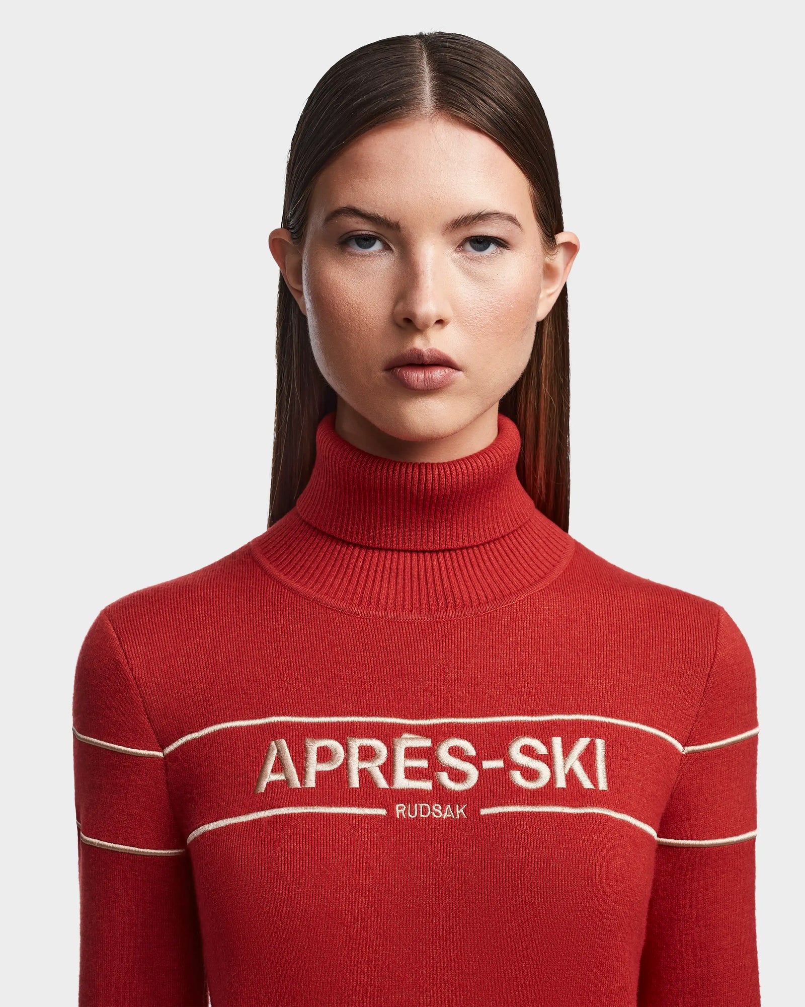 A woman with straight brown hair wears the ERIKA - 8523536 Orange après-ski turtleneck sweater from Rudsak, featuring bold white lettering. She faces forward against a light background—ideal for winter style.