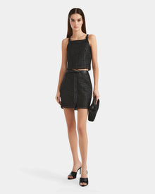 A woman faces forward in a black handwoven cropped top and matching skirt, styled with the Rudsak NADINE - 8524042 Black clutch and black open-toe heels against a white background—an ideal luxury wardrobe essential.