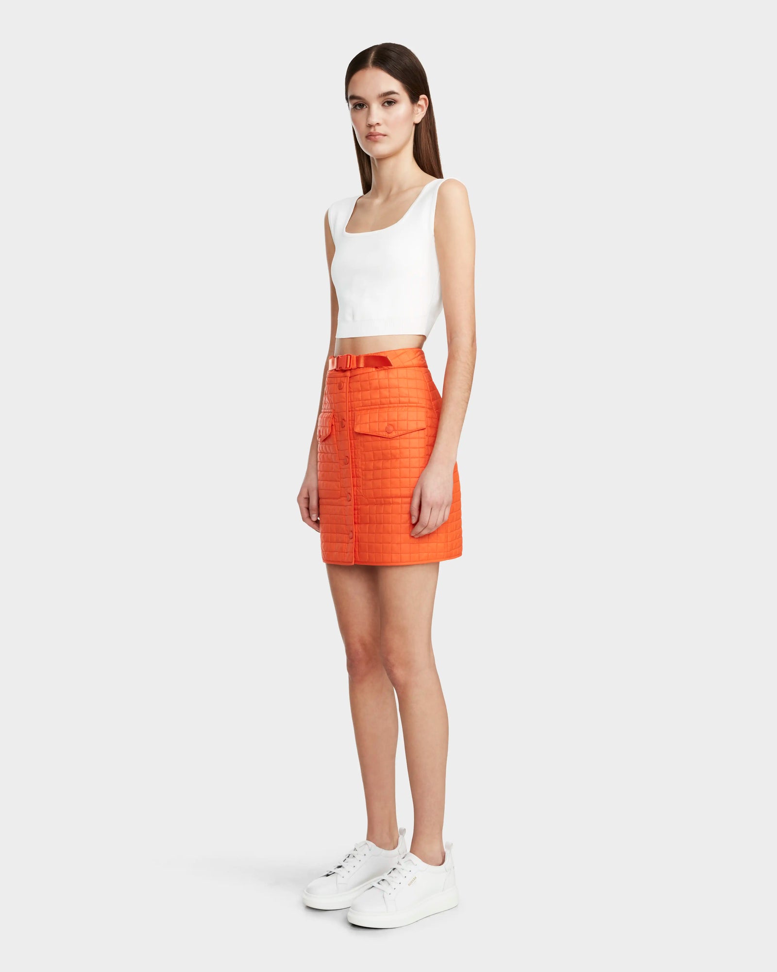 A woman faces to the side wearing Rudsaks SABINE - 8524063 Orange Sorbet quilted short skirt with large pockets, paired with a white sleeveless crop top and sneakers. A plain light gray background adds a modern touch.