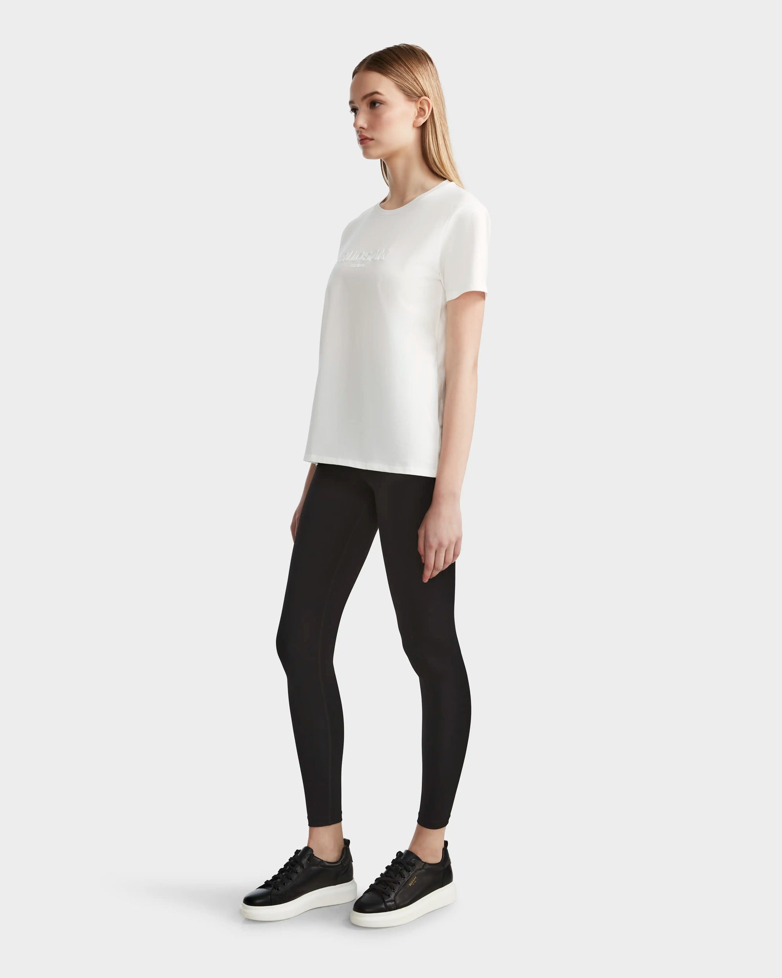 A woman stands against a plain background wearing the Rudsak AMINA - 8524012 Cotton White womens short sleeve t-shirt, black leggings, and black sneakers. She is facing right with her arms at her sides.