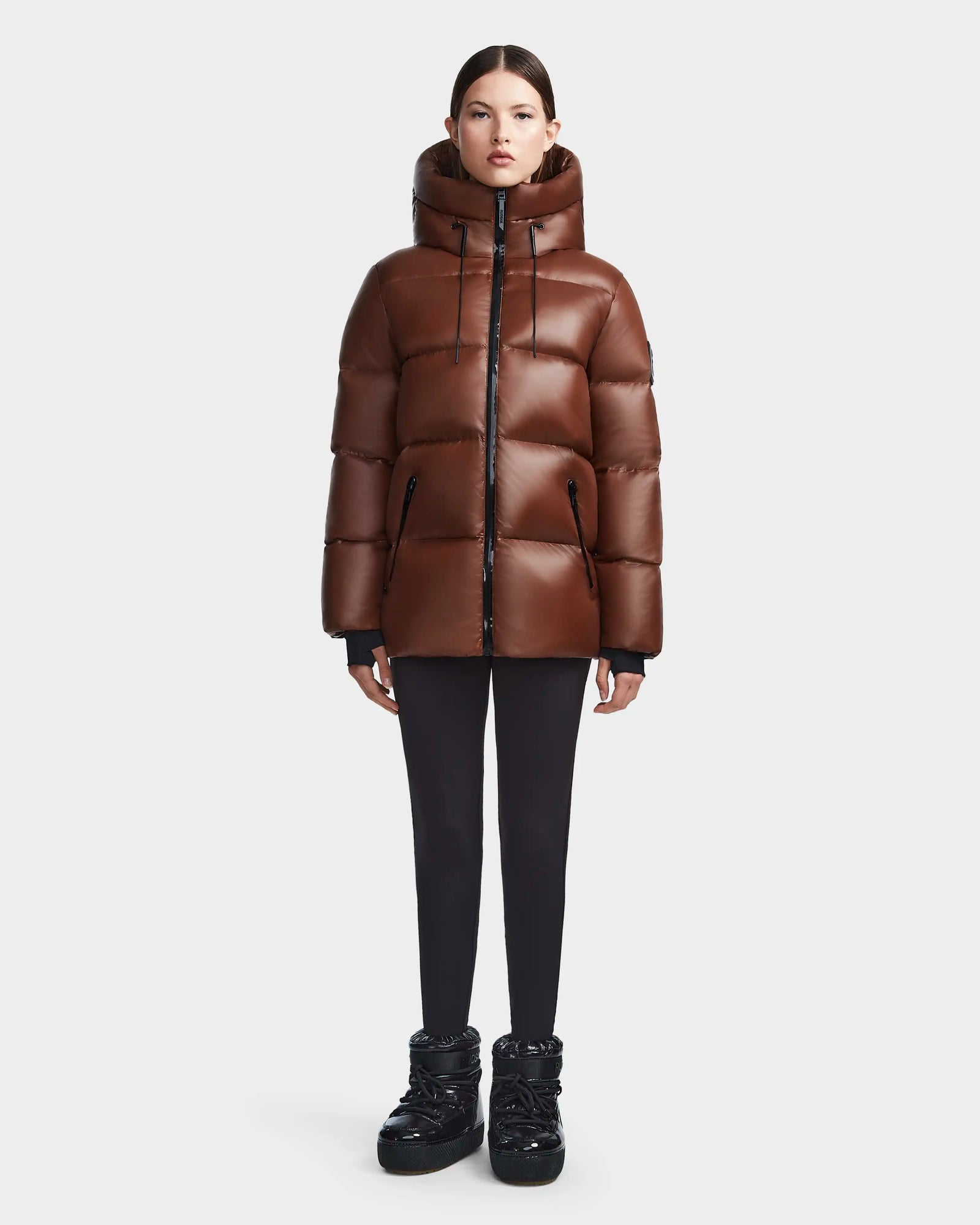A woman stands facing forward wearing the Rudsak RITA S - L123527 Cohiba shiny brown puffer jacket, black leggings, and black puffer boots against a plain light background.