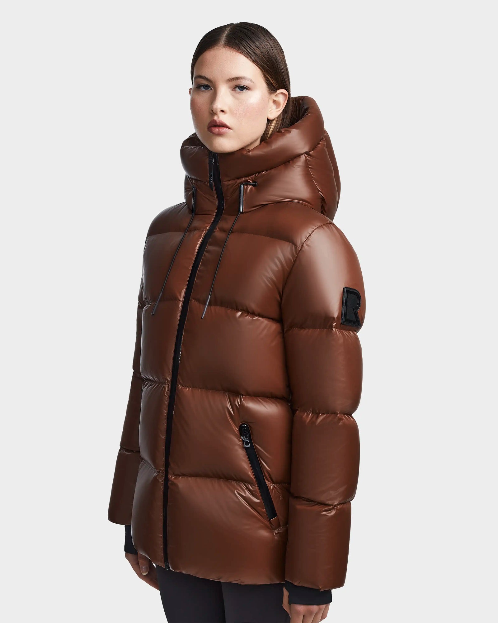 A woman stands against a light gray background wearing the Rudsak RITA S - L123527 Cohiba, a shiny brown weather-resistant puffer jacket with a high hood, black zippers, and an upper sleeve patch.