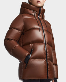 A person wears the Rudsak RITA S - L123527 Cohiba, a shiny brown weather-resistant puffer jacket with high collar, hood, black zippers, and drawstrings. The models face is partially visible, with one hand tucked into a pocket.