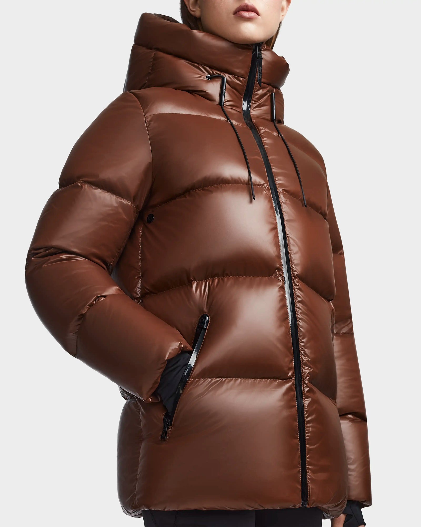 A person wears the Rudsak RITA S - L123527 Cohiba, a shiny brown weather-resistant puffer jacket with high collar, hood, black zippers, and drawstrings. The models face is partially visible, with one hand tucked into a pocket.