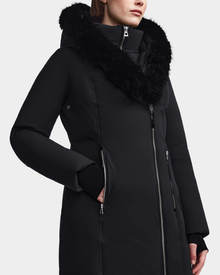 A person is wearing the Rudsak MAIA M - 8123582 Black womens down parka, featuring a large black faux fur hood and an asymmetrical front zipper. Only their lower face and upper body are visible.