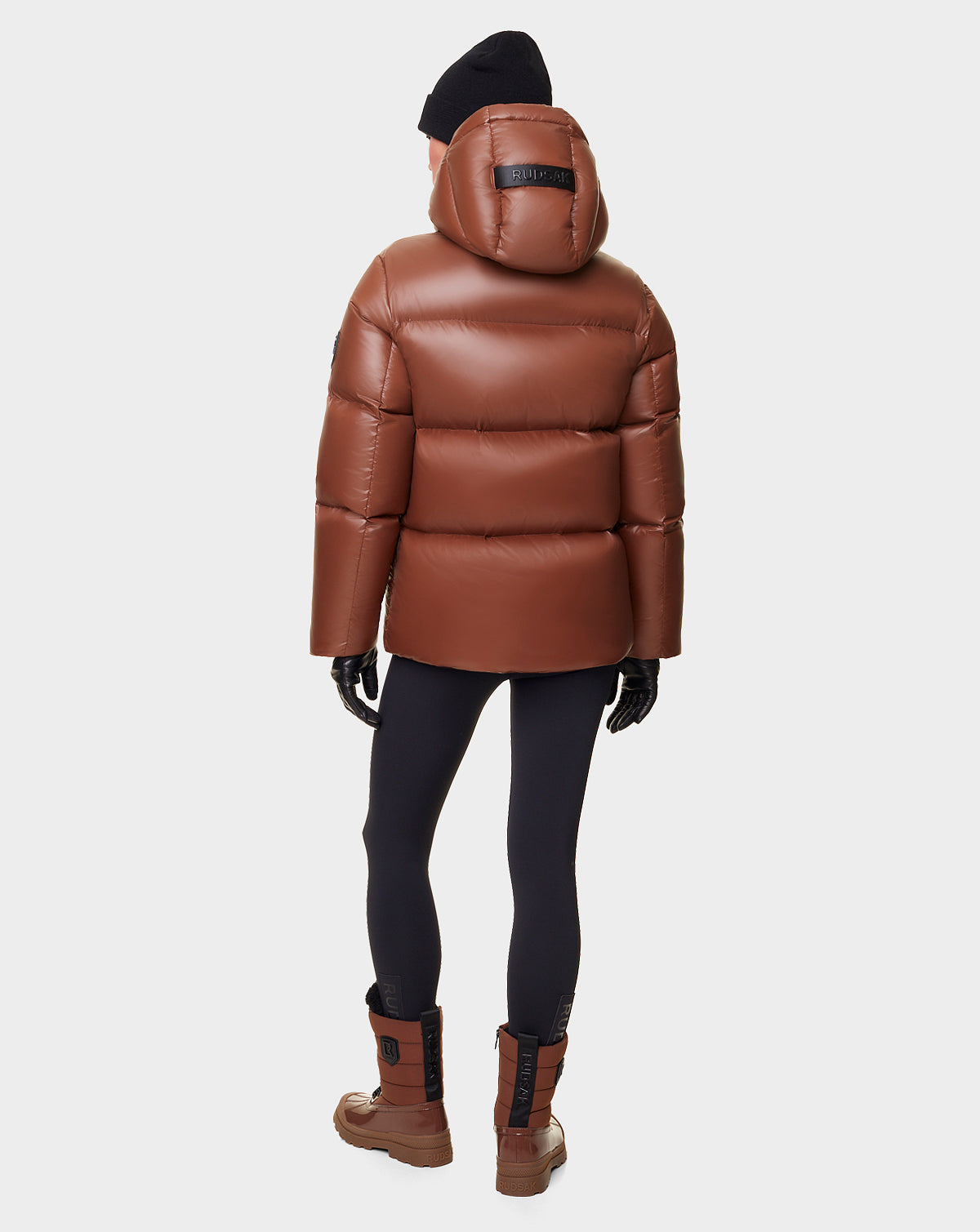 A person stands facing away against a white background, wearing the RUDSAK DORA - 8122547 COHIBA oversized brown down-filled puffer jacket with removable hood, paired with black leggings, brown boots, black gloves, and a black beanie.