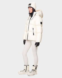 A woman wears the RUDSAK DORA - 8122547 ICELAND white down-filled puffer jacket with removable hood, styled with white leggings, winter boots, a beanie, and black gloves against a plain light background.