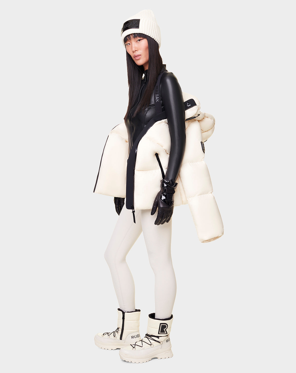 A woman wears the RUDSAK DORA - 8122547 ICELAND down-filled puffer jacket over a black top, white beanie, black gloves, white leggings, and chunky white boots against a plain light gray background.