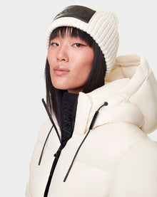 A woman with straight black hair and a neutral expression wears the DORA - 8122547 ICELAND white down-filled puffer jacket with a removable hood and a white knit beanie, both featuring black RUDSAK branding.