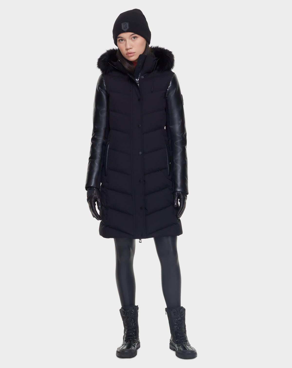 A woman faces forward wearing the Rudsak ELSA - L121519 BLACK sustainable winter coat with a fur-trimmed hood, black leather sleeves, leggings, lace-up boots, gloves, and a black beanie against a plain light background.