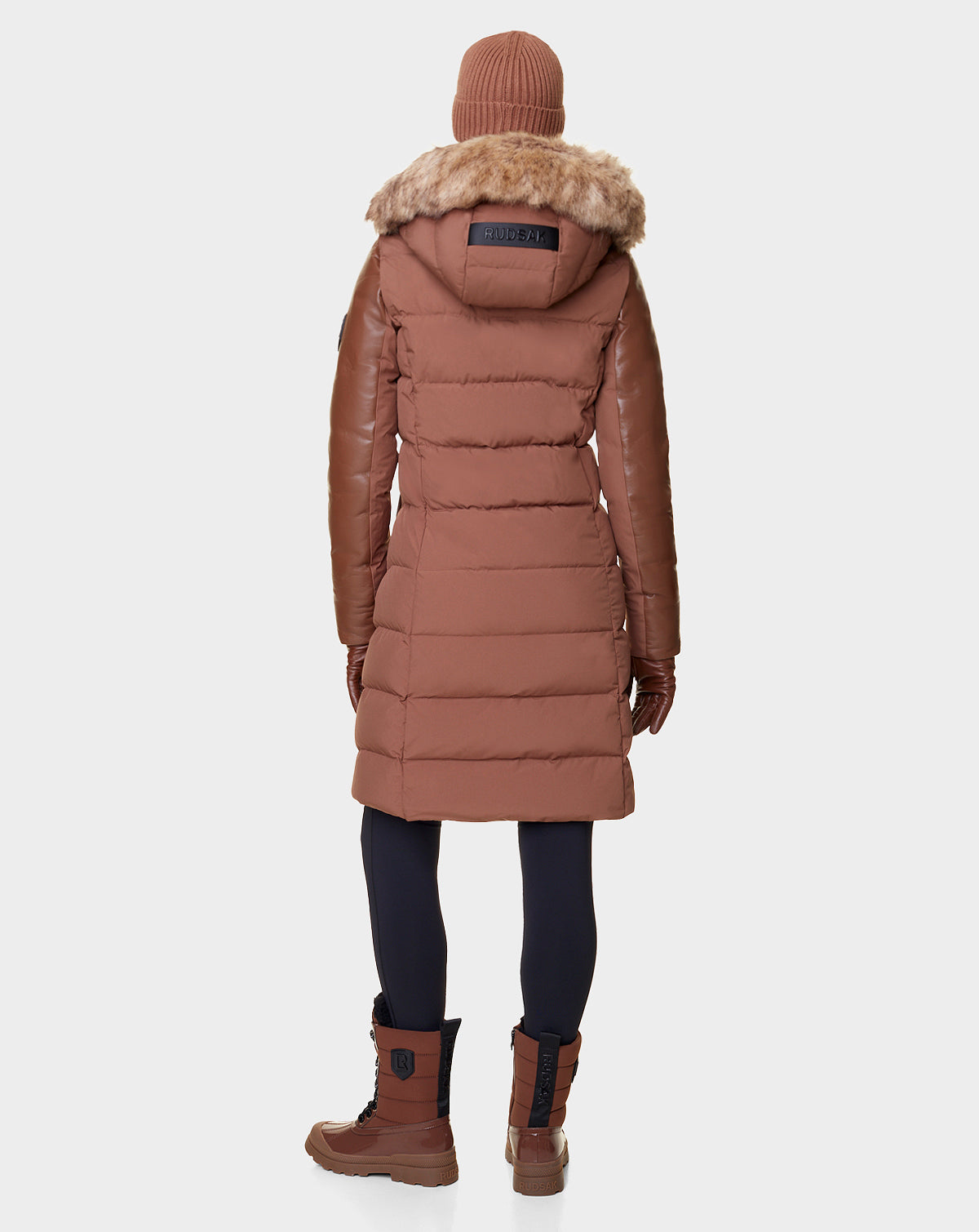 Wearing the Rudsak ELSA - L121519 TAUPE women’s quilted down puffer with a fur-trimmed hood, plus matching accessories and dark leggings, someone stands facing away against a plain background. The sustainable design features RDS-certified down.