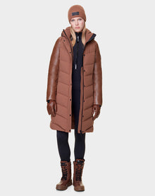 A woman stands facing forward in the Rudsak ELSA - L121519 TAUPE, a long taupe women’s quilted down puffer with leather sleeves—an elegant, sustainable look paired with brown gloves, knit beanie, black leggings, and brown winter boots.