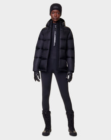 A person faces forward in a Rudsak EVIE - L122517 BLACK womens RDS-certified down puffer jacket, styled with black gloves, leggings, lace-up boots, and a beanie against a plain light gray background.