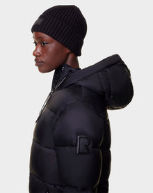 A person wears a black Rudsak EVIE - L122517 women’s down puffer with an R patch on the sleeve and a black beanie, looking left against a plain light background. The jacket features RDS-certified down for warmth and comfort.