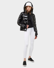 A woman faces forward against a plain light background, wearing the MARE - 8121011 BLACK women's down puffer jacket with a hood by Excluded, paired with a white baseball cap, black top, white fitted pants, and black sneakers.