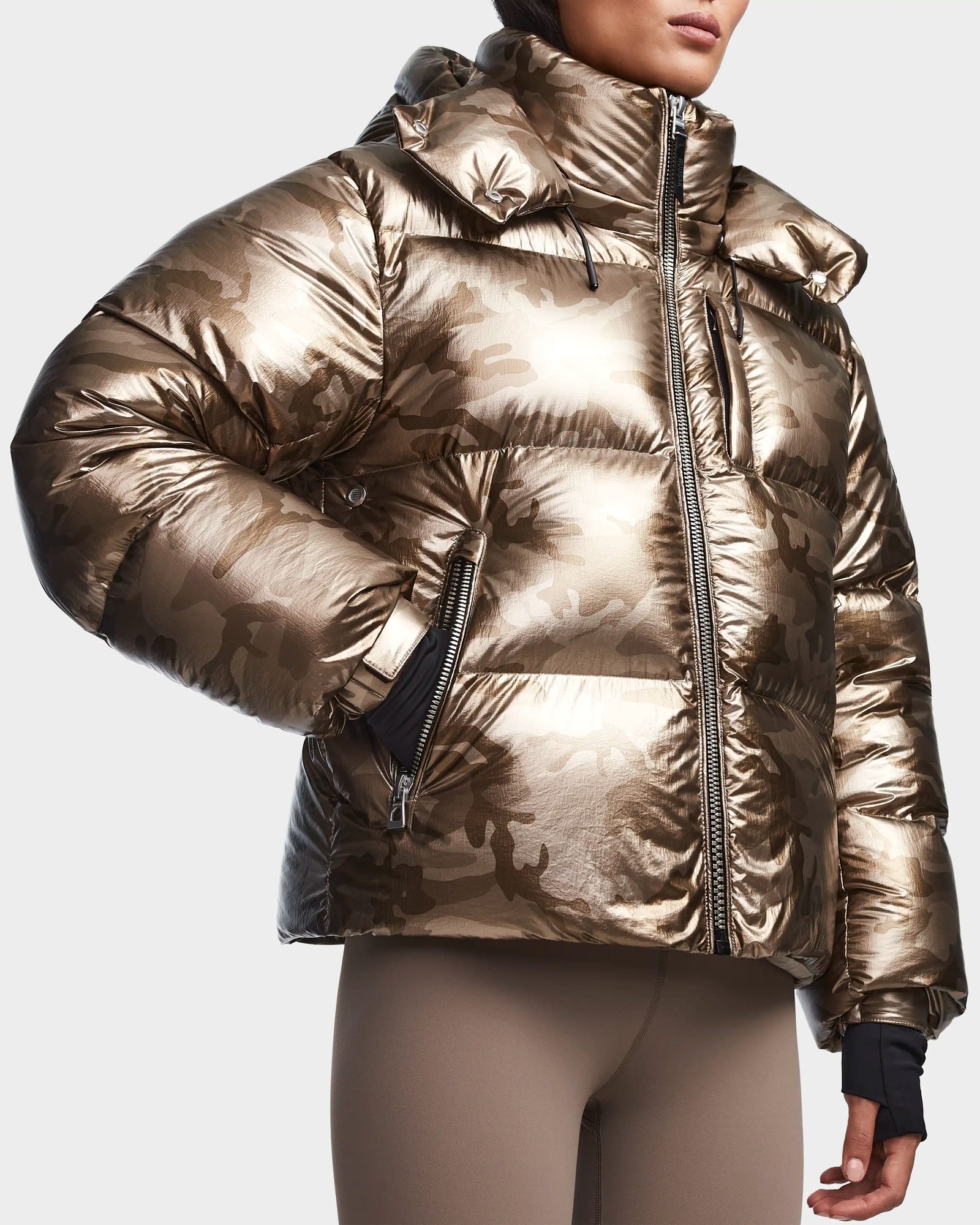 A person wearing the Rudsak MAVE S - 8123563 Metallic Ivory Camo womens down puffer with a hooded front zipper and lightweight insulation, paired with light brown leggings. The water-repellent jacket is shown from the shoulders down.