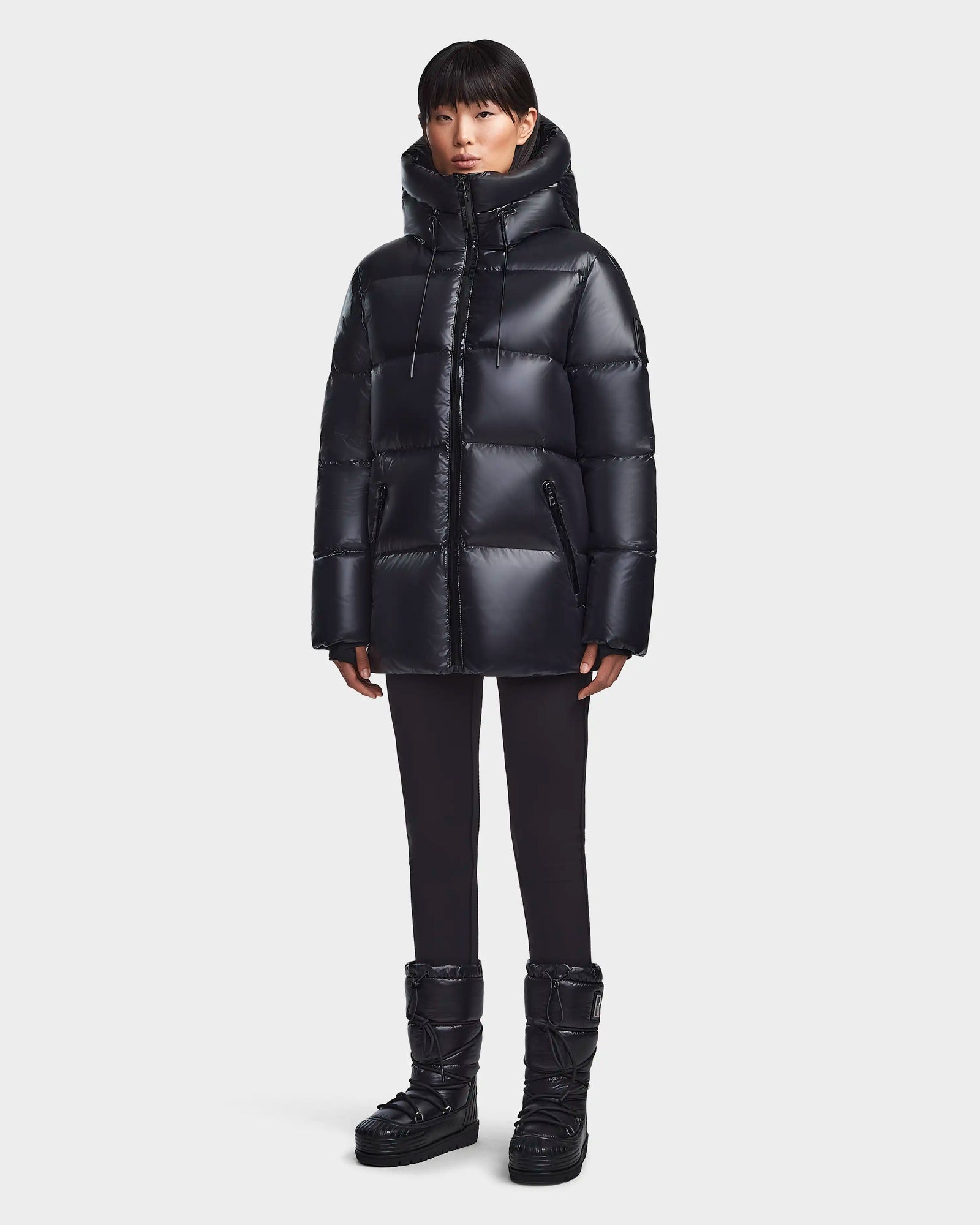 A person models the Rudsak RITA S - L123527 Black women’s down puffer jacket with matching black pants and black puffer boots against a plain light gray background.