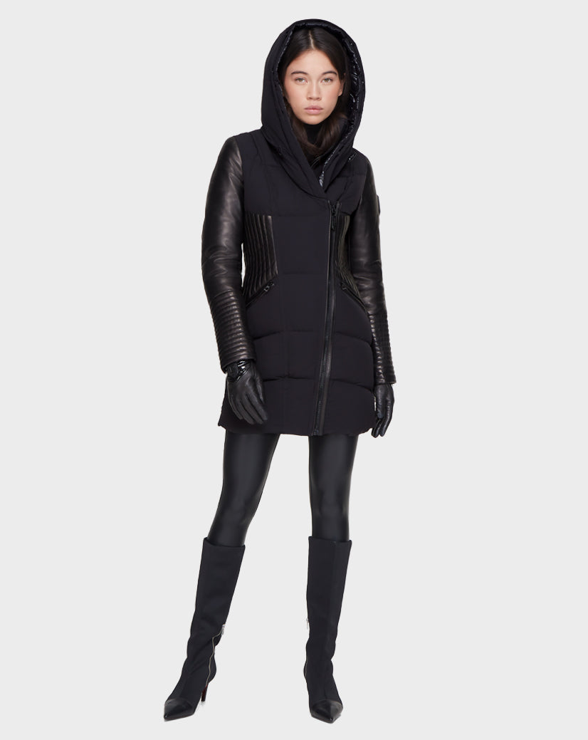 A woman faces forward, wearing the Rudsak SHAUNA - 8121509 BLACK winter coat with leather sleeves, paired with black gloves, leggings, and knee-high heeled boots against a plain light background.
