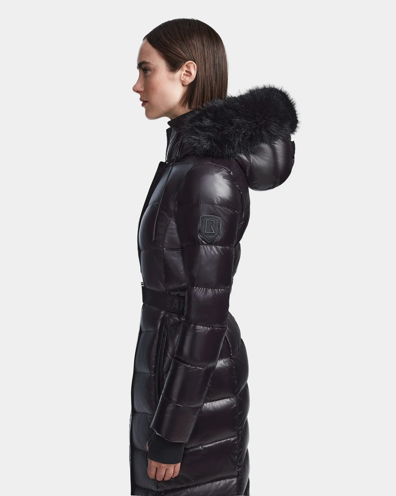 A woman stands in profile wearing the Rudsak ELSIE S - L123518 Black, a long black maxi down puffer coat with a belted waist and faux fur-trimmed hood, set against a plain white background.