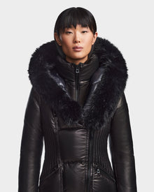 A person wears the SHAUNA X - 8121568 BLACK women's winter coat by Rudsak, featuring cloud leather and a large fur-trimmed hood, standing against a plain white background and looking straight ahead with a neutral expression.