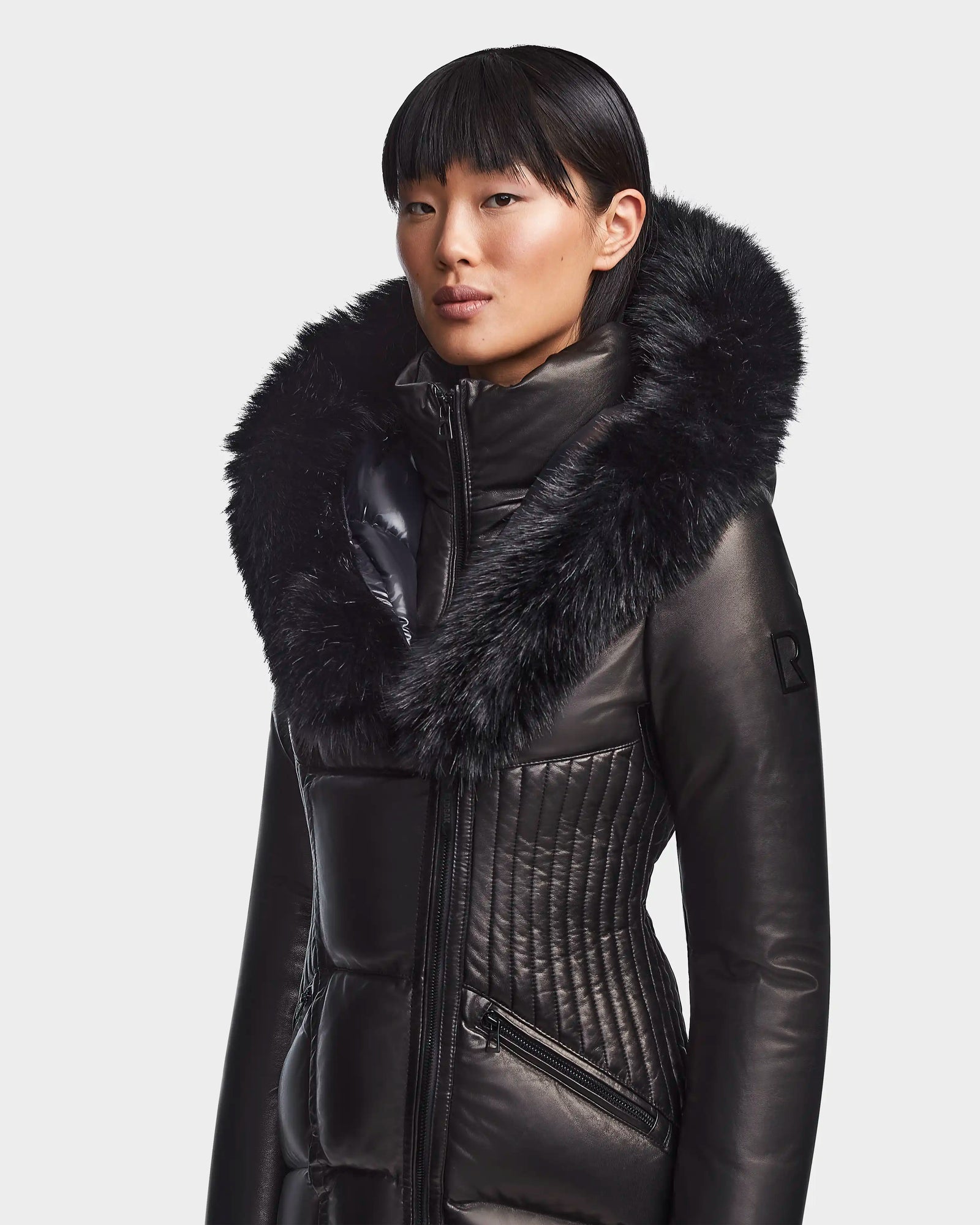 A person wears the Rudsak SHAUNA X - 8121568 BLACK women's winter coat, featuring a sleek design and a large faux fur-trimmed hood, standing against a plain light background.