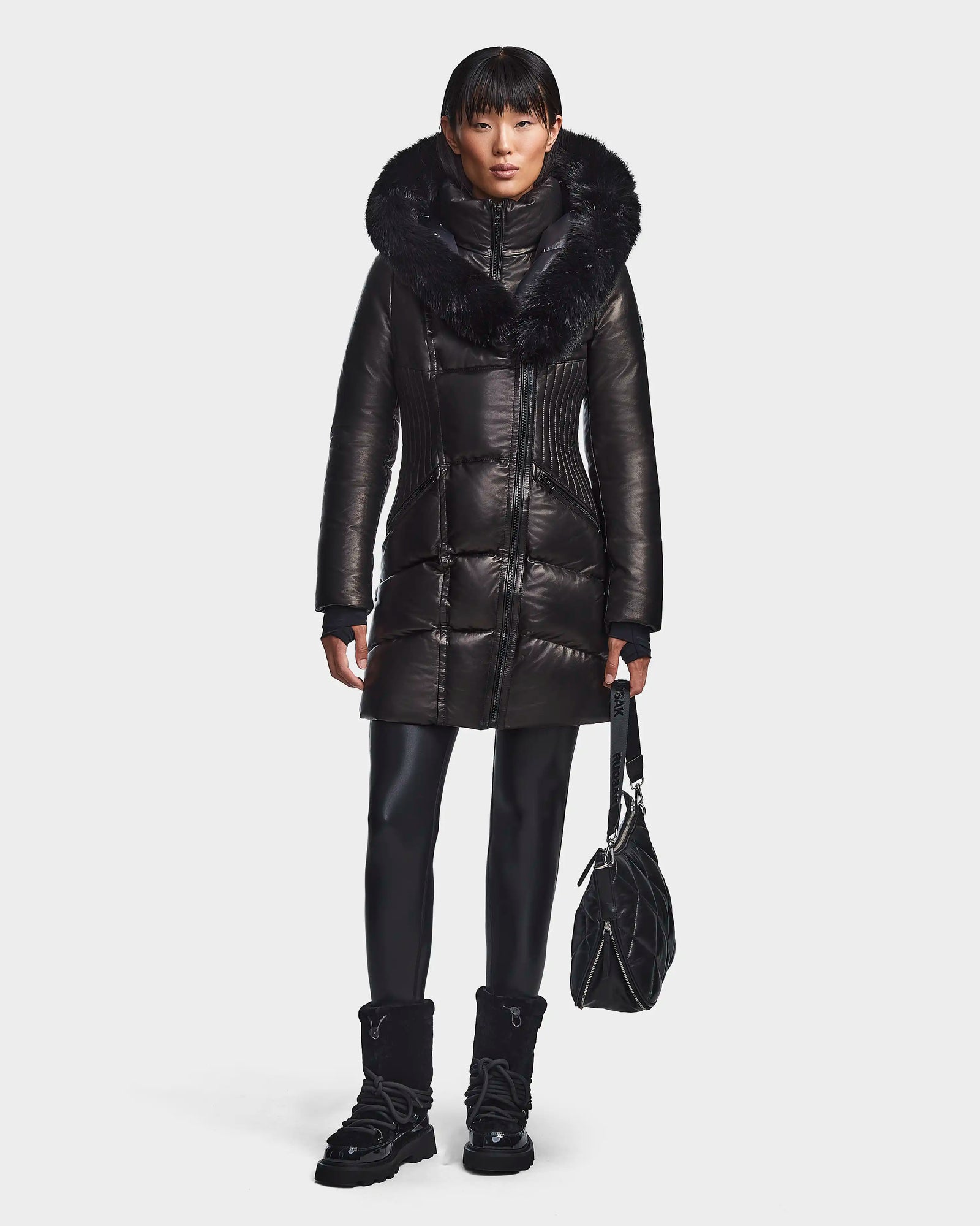 A person faces forward against a white background, wearing the Rudsak SHAUNA X - 8121568 BLACK women's winter coat with a large fur-trimmed hood and RDS down, paired with black leggings, boots, and holding a black quilted bag.