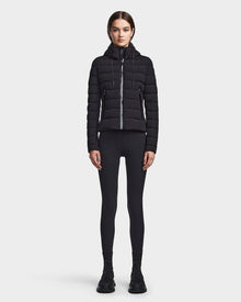 A woman faces forward against a white background, wearing the RUDSAK MELISSA - 8122551 BLACK womens puffer jacket with a hood, along with black leggings and black sneakers.