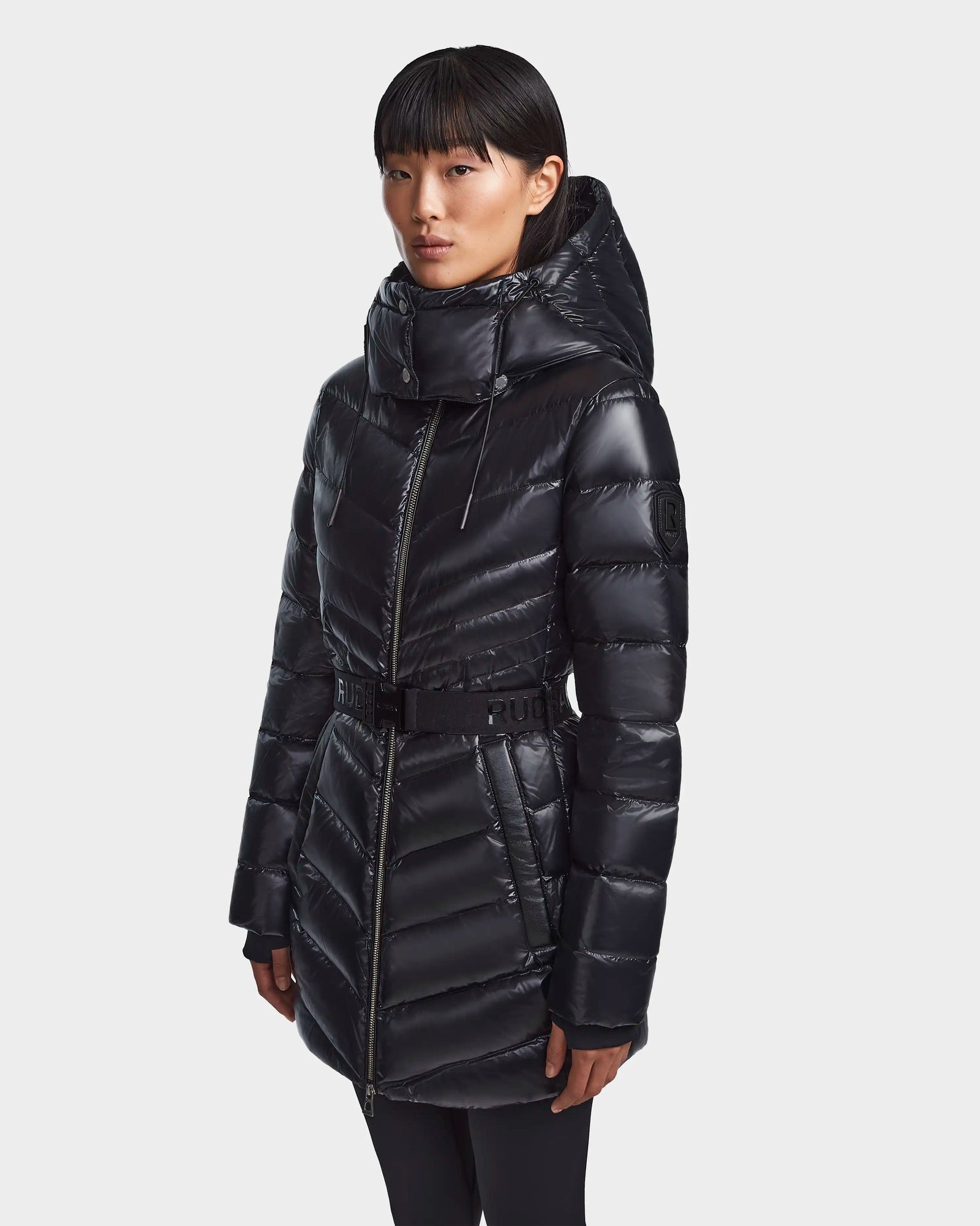 A woman wears the Rudsak MALAYA - L123510 Black down puffer jacket with herringbone quilting, high collar, hood, and belted waist, standing against a plain light background.