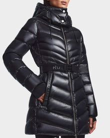 A woman wears Rudsak’s MALAYA - L123510 Black, a recycled glossy black down puffer jacket for women with herringbone quilting, hood, matching belt with RUD logo, zip-up front, and side pockets.