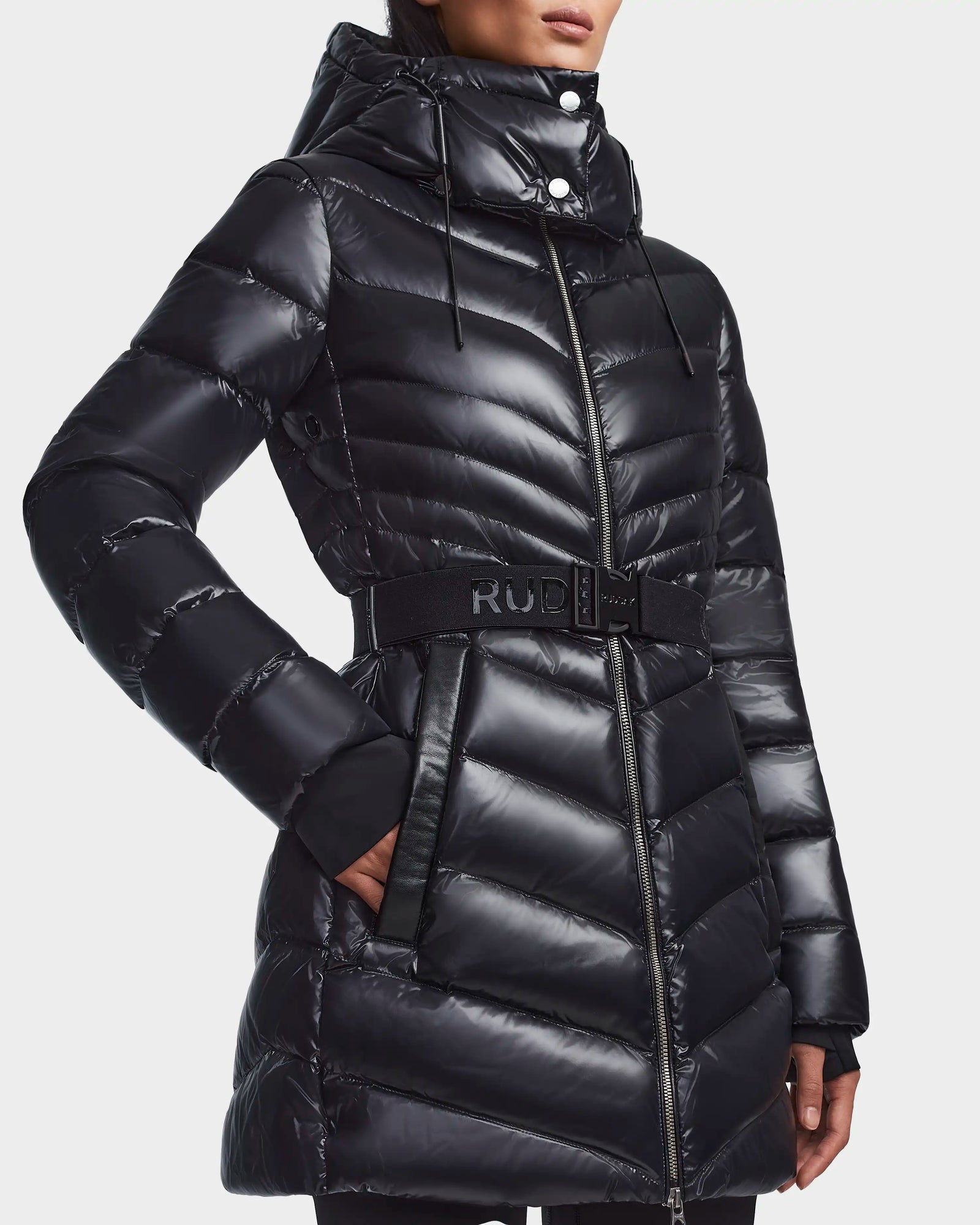 A woman wears Rudsak’s MALAYA - L123510 Black, a recycled glossy black down puffer jacket for women with herringbone quilting, hood, matching belt with RUD logo, zip-up front, and side pockets.