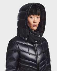 A person with straight black hair and bangs models the Rudsak MALAYA - L123510 Black recycled glossy women’s down puffer jacket with herringbone quilting, large hood up and zipped to the neck, against a plain white background.