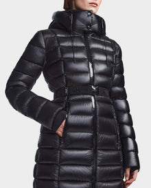 A person wearing the Rudsak FRIDA S - L123513 Black, a women's mid-length, glossy quilted down jacket with a removable hood and matching belt. Hands are in pockets; face is partially out of frame.