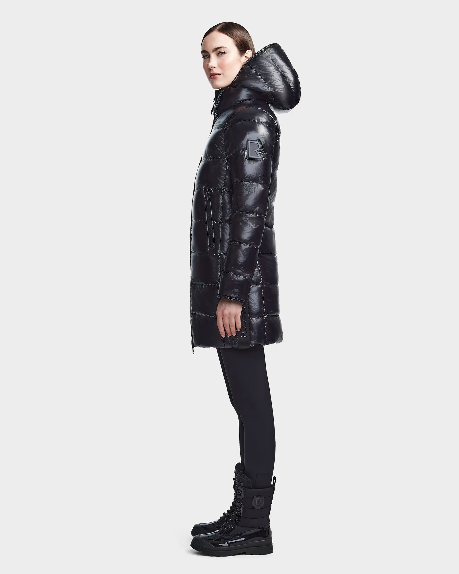 A woman wears the RUDSAK MARNIE LONG - 8122519 BLACK puffer jacket with a hood, paired with black leggings and winter boots, standing in profile against a plain light background.