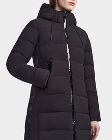 A woman stands against a plain background wearing the RUDSAK MARNIE LONG - 8122519 BLACK jacket, which features a hood, shiny black zipper and trim, and has one hand in a side pocket.