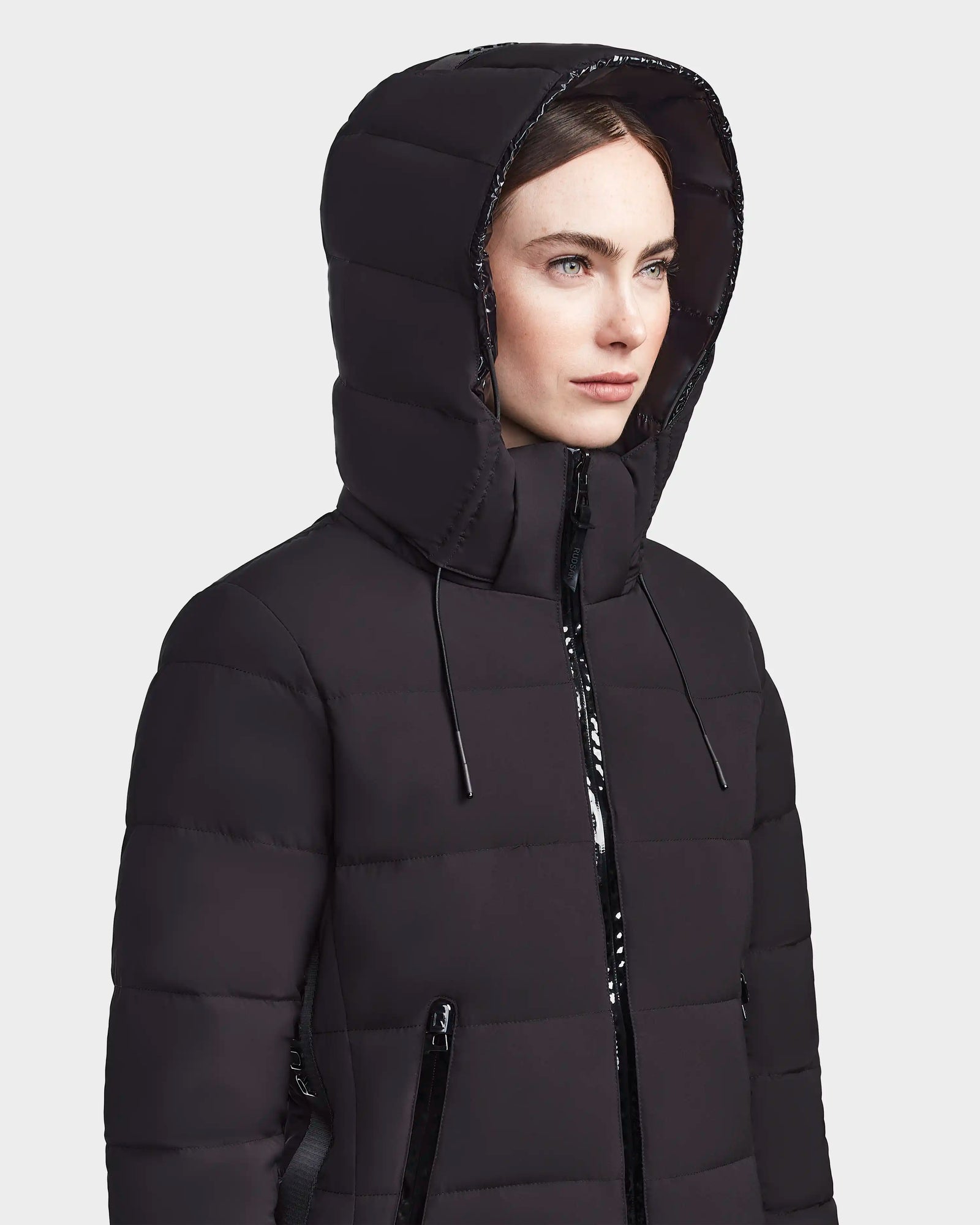A woman wears the RUDSAK MARNIE LONG - 8122519 BLACK jacket with a hood, front zipper pockets, and drawstrings, looking off to the side against a plain light background.