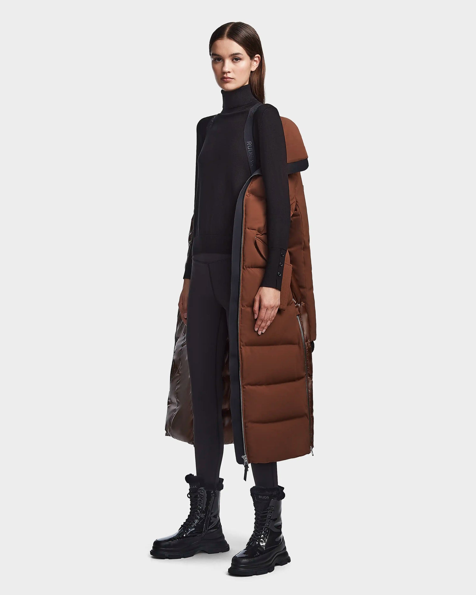 A woman models the RUDSAK CALI - 8122502 COHIBA long brown puffer coat over one shoulder, styled with a black turtleneck, black leggings, and lace-up boots against a plain white background.
