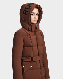 A woman wears the CALI - 8122502 COHIBA maxi puffer coat by RUDSAK, featuring a brown belted design, large hood with bold RUD print, RDS-certified down filling, zip and snap closures, and black drawstrings.