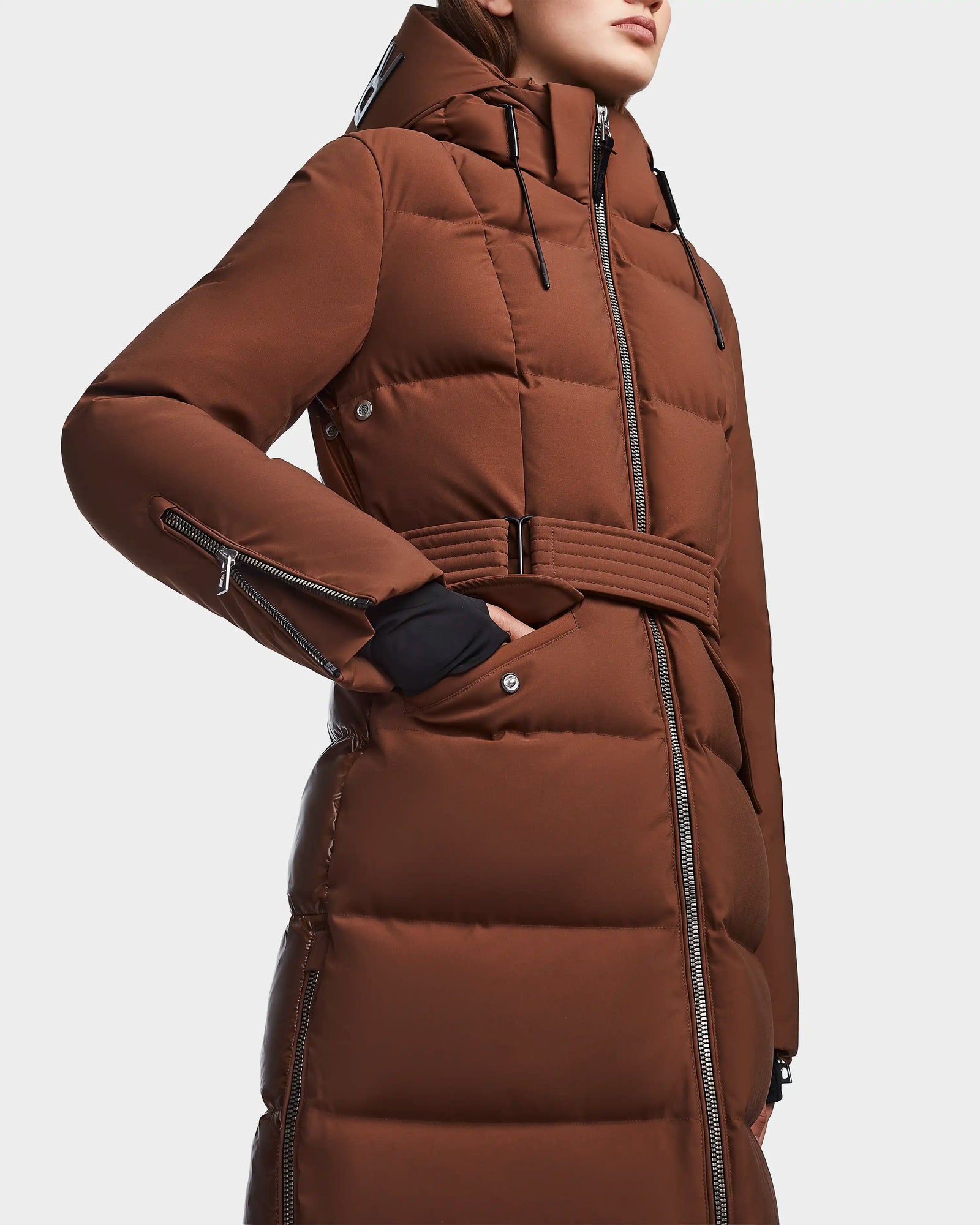 A person wearing the RUDSAK CALI - 8122502 COHIBA women’s brown belted winter coat with RDS-certified down, a hood, and zippered details poses with one hand in the pocket against a plain background.