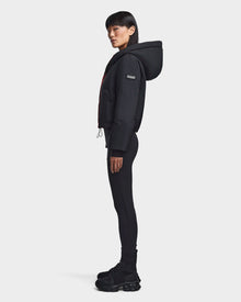 A person stands in profile against a plain background, wearing the Rudsak BALA - 8123614 Orange women’s hooded puffer jacket, paired with black leggings and chunky sneakers.