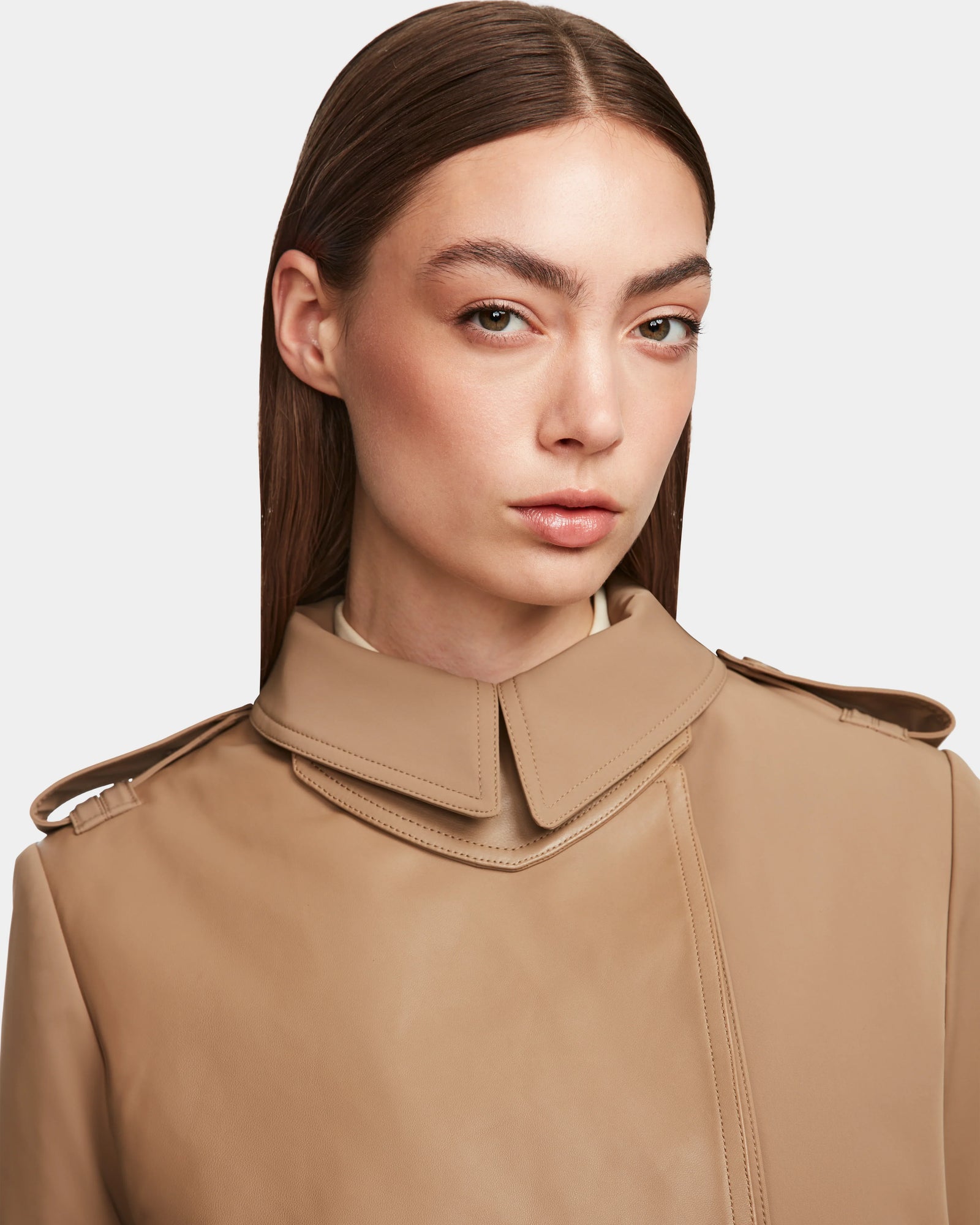 A woman with straight brown hair and natural makeup models the Rudsak KINDA - 8124690 Camel womens trench coat, featuring a unique double-layered collar, as she looks confidently at the camera against a plain light background.