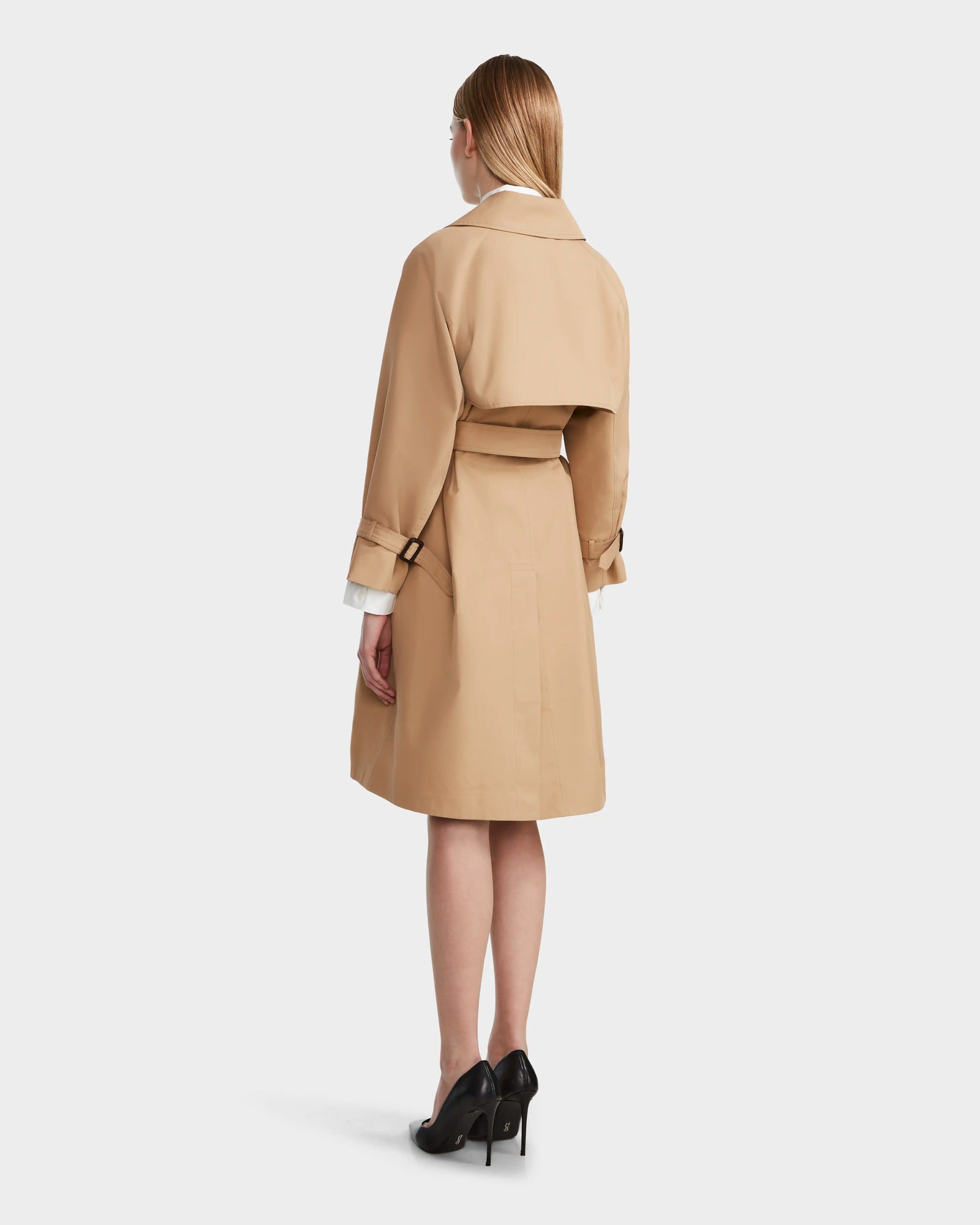 A woman with straight blonde hair faces away, wearing the Rudsak MIRELLE - 8124055 Khaki womens trench coat and black high heels against a plain white background.