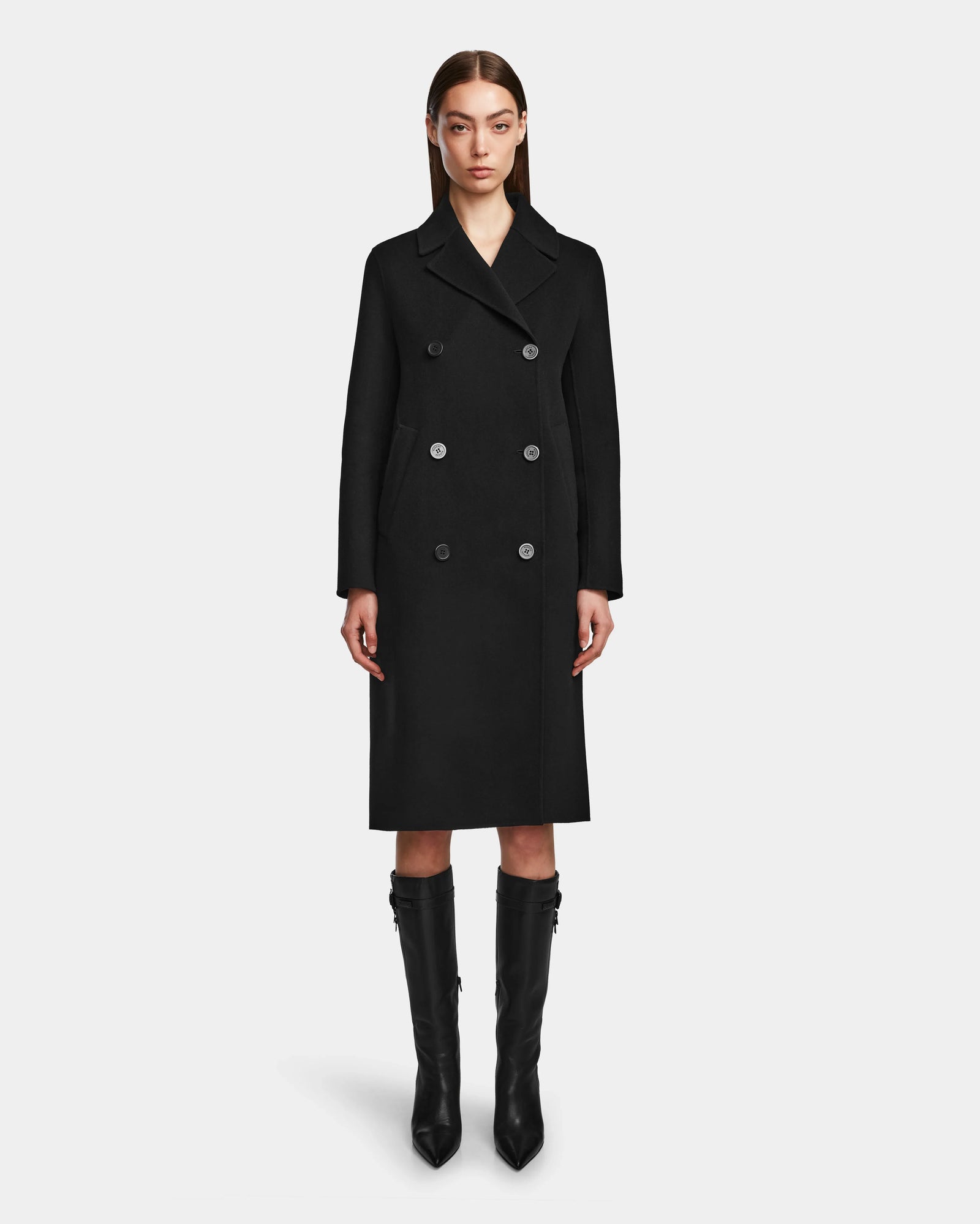 A woman with straight brown hair wears the Rudsak LAYLAH - 8124699 Black knee-length double-breasted women’s wool coat and knee-high black boots, standing against a plain white background.