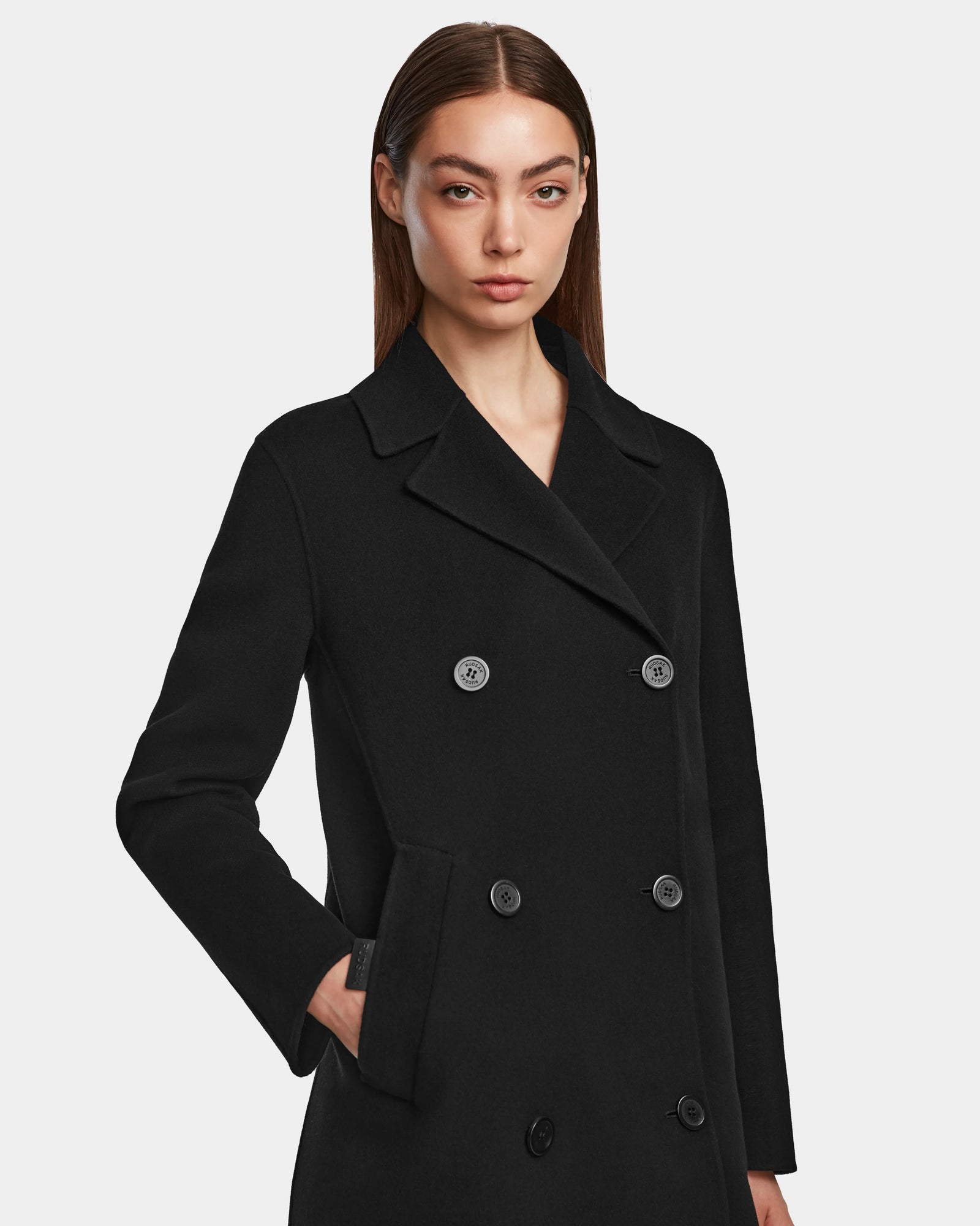 A woman with straight brown hair wears the Rudsak LAYLAH - 8124699 Black womens wool coat, double-breasted and knee-length, her left hand in a pocket, standing against a plain light background with a neutral expression.