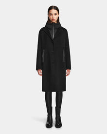 A woman faces forward against a white background, wearing Rudsak’s ARLA - 8124579 Black wool coat with leather sleeves, paired with black leggings, a black zip-up underneath, and black boots.