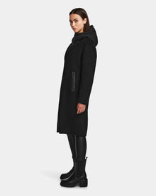 A woman stands confidently in the Rudsak ARLA - 8124579 Black coat, its classic double-breasted design elevated by sleek leather sleeves.
