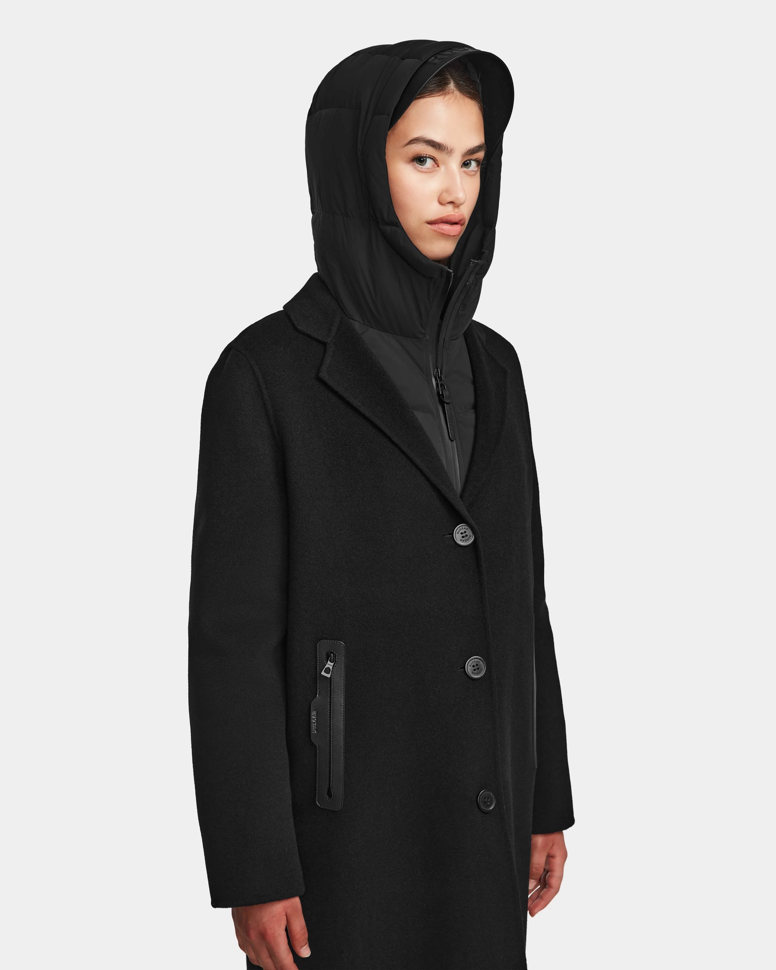 A woman wearing the Rudsak ARLA - 8124579 Black womens wool coat with leather sleeves and a zipper pocket stands against a plain light gray background, looking slightly to the side with a neutral expression.