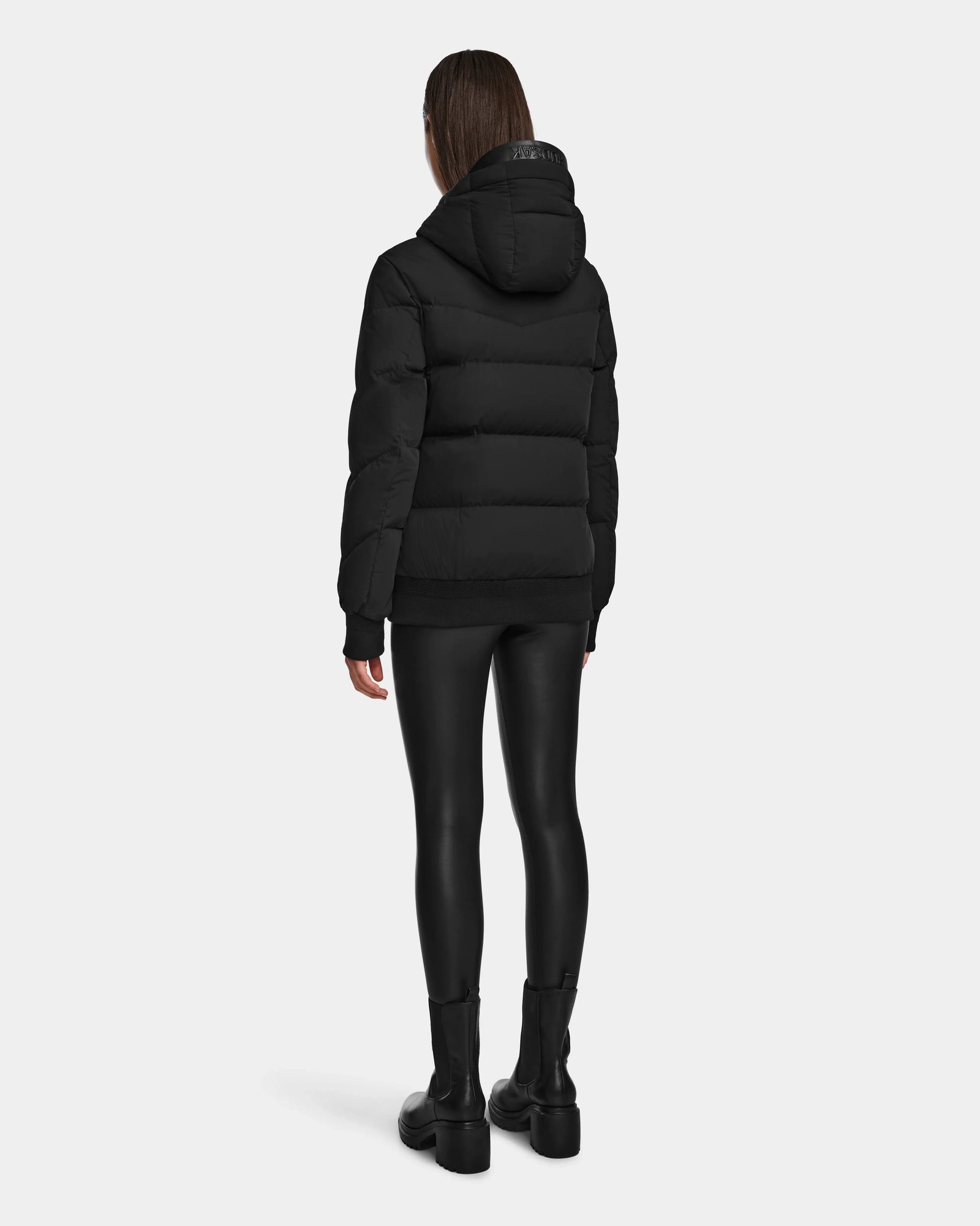 A person stands facing away against a plain white background, wearing the Rudsak ARLA - 8124579 Black hooded puffer jacket with leather sleeves, paired with black leggings and black boots.