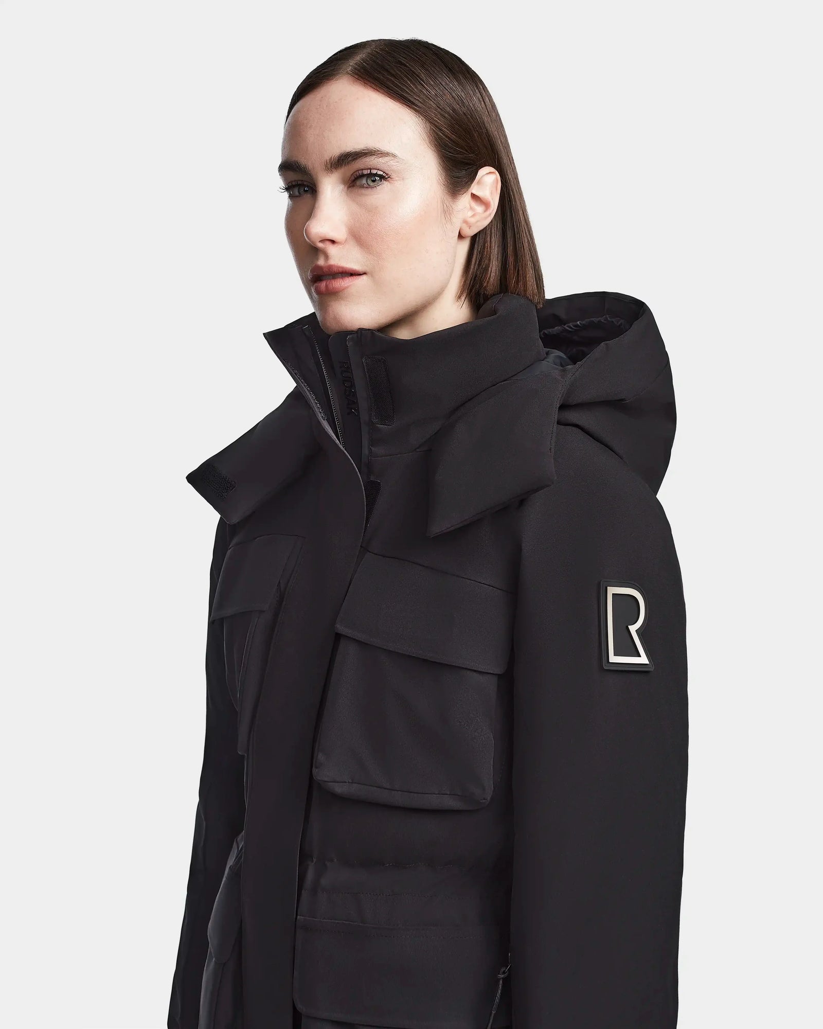 A woman with straight brown hair wears the Rudsak LUX - 8123618 Black ski jacket, featuring a hood, large front pockets, and an R patch on the upper sleeve. She looks to the side against a plain, light background, showcasing winter style.