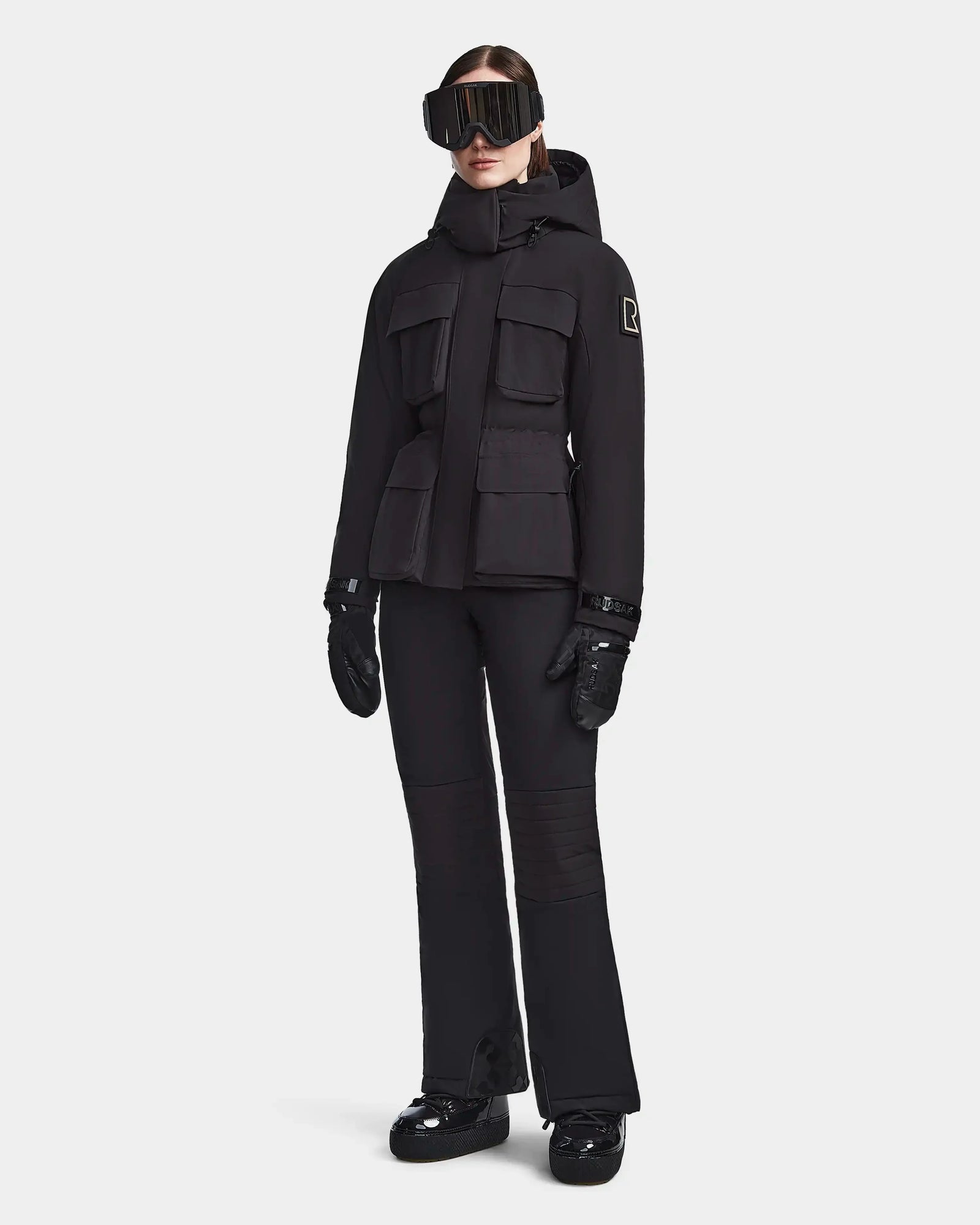 A person stands facing forward against a plain white background, dressed in the Rudsak LUX - 8123618 Black women’s ski jacket, paired with matching ski pants, gloves, boots, and large ski goggles for a sleek winter look.